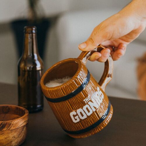 Primer plano de mano sosteniendo la jarra 3D de The Goonies con diseño de barril pirata, llena de bebida espumosa.