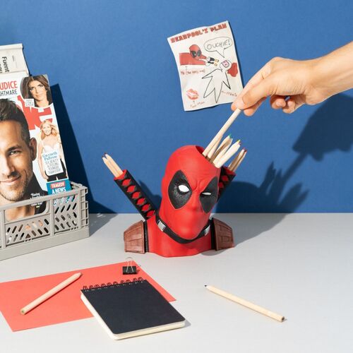 Figura de Deadpool en un escritorio azul y gris, con lápices insertados en la cabeza y las vainas de la espalda, en un entorno de oficina.